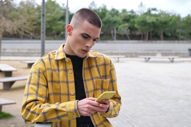 Handsome young man wearing make up, chatting on his smartphone annoyed. Non binary androgynous guy. Handsome young man wearing make up, chatting on his smartphone annoyed. Non binary androgynous guy. sad gay stock pictures, royalty-free photos & images