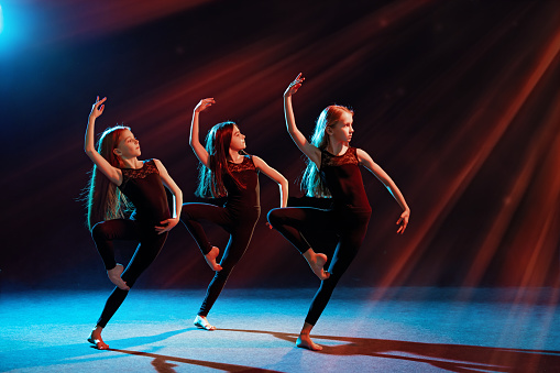 group of three ballet girls in tight-fitting costumes dance against a black background with their long hair down, silhouettes illuminated by color sources.