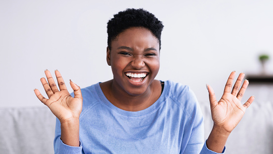Portrait of excited emotional young curvy African American woman smiling and laughing looking at camera while sitting on the sofa at home in living room. Cheerful chubby lady posing, hearing good news
