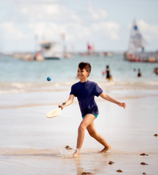 Smiling young boy having fun at the beach in summer Smiling boy running along a sandy beach with a paddle and ball in the summer paddle ball stock pictures, royalty-free photos & images