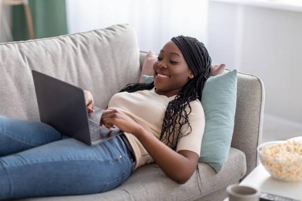 istock Joyful african lady laying on couch at home, using laptop 1300316406