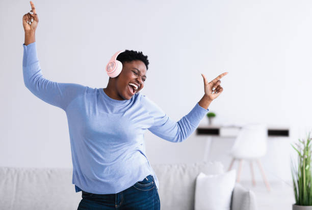 Black woman listening to music in headphones and dancing Enjoying Favorite Song. Emotional excited curvy black woman wearing wireless headset listening to music, dancing in living room with closed eyes and raising hands up. Positive lady having fun huge black woman pictures stock pictures, royalty-free photos & images