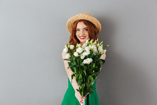 Photo of charming teenage woman with red hair wearing straw hat looking on camera and holding bunch of white flowers lisianthus isolated over gray wall