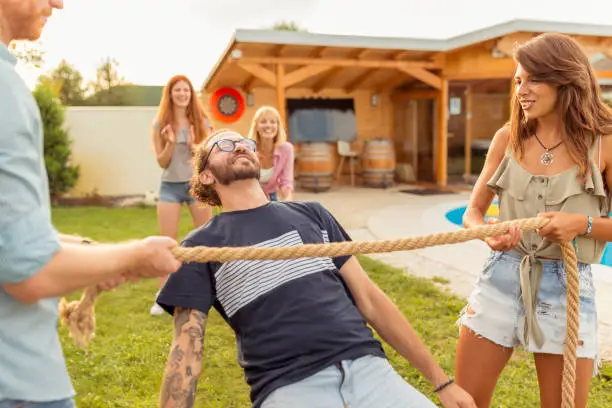 Group of cheerful young friends having fun at summertime outdoor party by the swimming pool, participating in limbo dance contest, passing below the rope while dancing