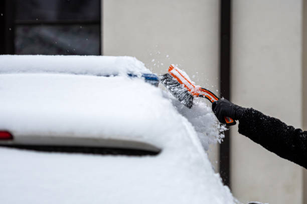 femmina bionda in una maschera facciale con uno squittio pulisce la neve da un'auto parcheggiata nel cortile - snow car window ice scraper foto e immagini stock