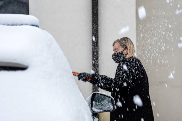 femmina bionda in una maschera facciale con uno squittio pulisce la neve da un'auto parcheggiata nel cortile - snow car window ice scraper foto e immagini stock