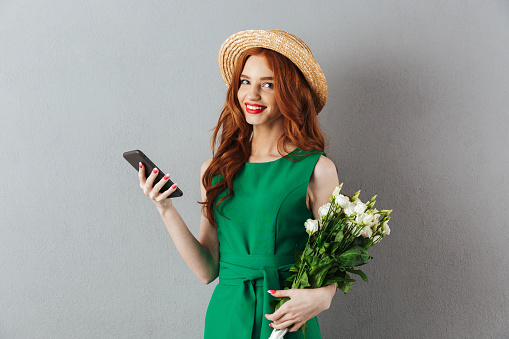 Image of redhead young happy woman in green dress and hat standing over grey wall background looking camera with flowers chatting by mobile phone.