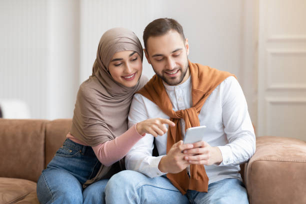 istock Modern Muslim Couple Using Mobile Phone Sitting On Couch Indoors 1300323274