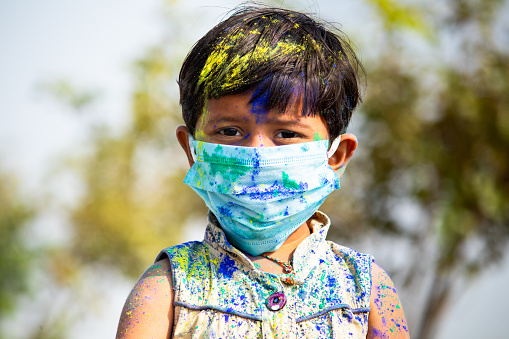 Portrait of young cute little girl kid with medical face mask with applied holi powder looking at camera - concept of holi celebration during coronavirus or covid-19 pandemic with safety measures