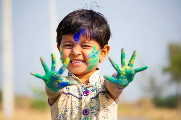 Photo of young cute cheerful little girl kid with applied holi colors powder showing colorful hands to camera during holi festival celebration.