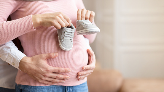 Pregnancy. Unrecognizable Couple Awaiting Baby, Husband Holding Tiny Shoes Near Pregnant Wife's Belly Posing Standing Indoor. Parents-To-Be, Family Moments, New Life Concept. Cropped