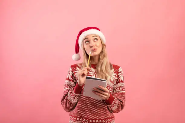 Photo of Charming young lady in Christmas outfit thinking about her New Year resolutions, writing wish list over pink background