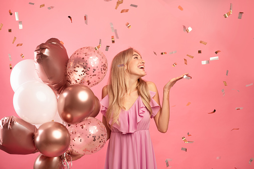 Charming young lady holding balloons, having birthday party over pink studio background with falling confetti, free space. Beautiful millennial woman celebrating holiday. Festive concept