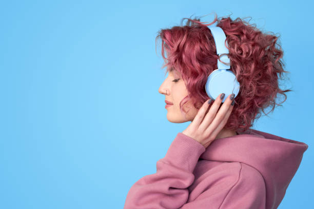 Young pink hair girl listening music in headphones Profile view portrait of dreamy young pink hair girl wearing in casual clothes, enjoying sound and listening music in headphones standing against blue background with copy space headphones stock pictures, royalty-free photos & images