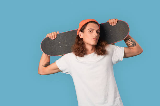 istock Freestyle. Curly-haired guy wearing hat standing isolated on blue with skateboard on shoulders posing confident 1300324599