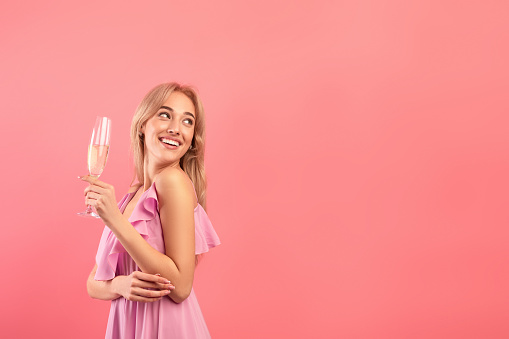 Beautiful blonde woman in dress holding glass of sparkly champagne, celebrating holiday, looking aside at empty space over pink studio background. Festive concept