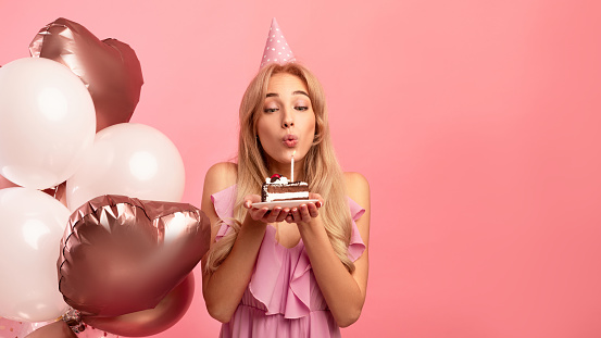 Cute young lady with birthday hat and balloons blowing candle on piece of cake over pink studio background, panorama with copy space. Millennial woman having holiday party or celebration