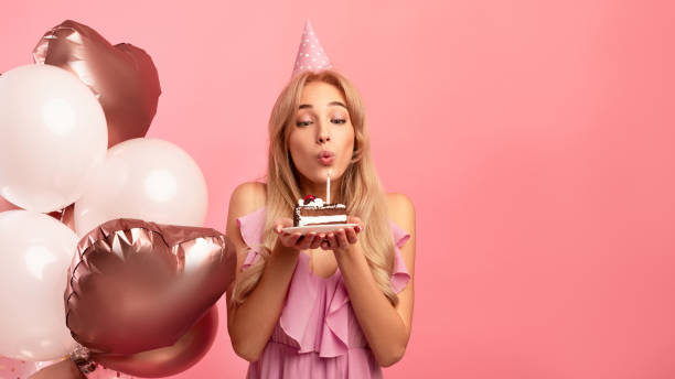 Cute young lady with birthday hat and balloons blowing candle on piece of cake over pink background, copy space Cute young lady with birthday hat and balloons blowing candle on piece of cake over pink studio background, panorama with copy space. Millennial woman having holiday party or celebration woman birthday cake stock pictures, royalty-free photos & images