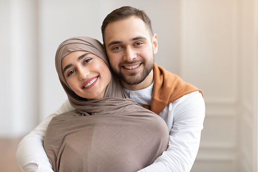 Cheerful Young Muslim Couple Hugging Posing Smiling To Camera Standing Indoors At Home. Arab Spouses In Love. Happy Arabic Family Portrait. Happiness In Marriage And Romantic Relationship Concept