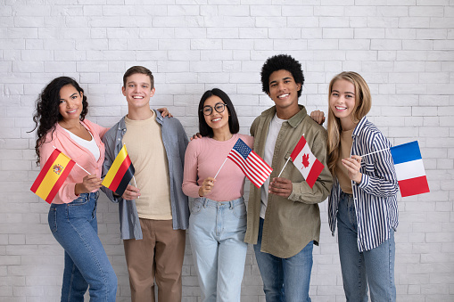 Immigrants of different countries and exchange students in language school. Smiling multiracial young people hold flags of France, Canada, USA, Germany and Spanish in classroom, studio shot