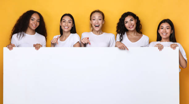 istock Multicultural Ladies Holding Empty Paper Board Over Yellow Background, Mockup 1300328232