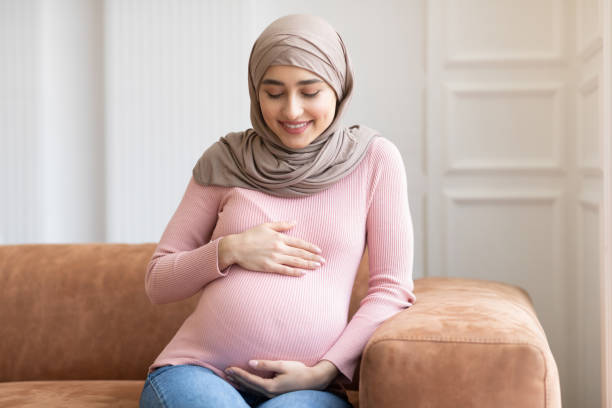 istock Pregnant Muslim Lady In Hijab Posing Sitting On Couch Indoor 1300328310