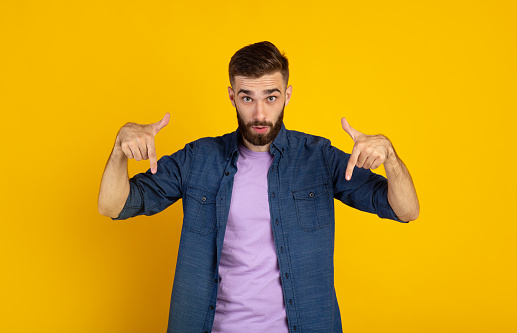 Surprised bearded guy pointing downwards on orange studio background, panorama. Emotional Caucasian guy showing or advertising something, offering big sale or product promotion