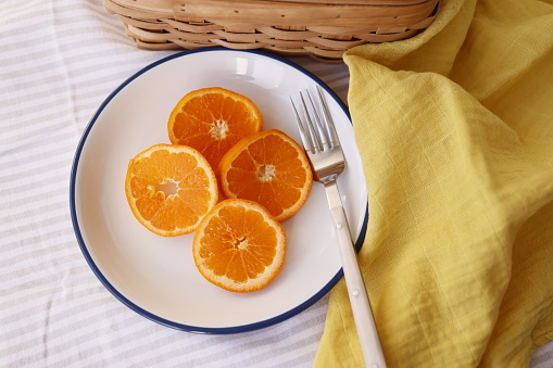 Istanbul, Turkey-February 3, 2021: On a White-Gray Striped Fabric Floor, Four Sliced Tangerine Slices in a White Ceramic Plate, The rim of the plate is dyed dark blue, the plate has a white bone handle fork, next to the plate is a yellow cloth napkin. Full Frame, Still life, Studio shot, Flat lay. Canon EOS R5, Canon RF 35mm Lens.