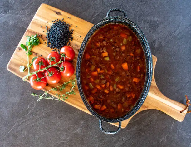 fresh cooked spicy minced meat stew with lentils and mediterranean vegetables served in a stew pot on wooden board from above on dark table background