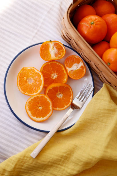 istock On a White-Gray Striped Fabric Floor, A White Ceramic Plate, Sliced Tangerines in a Ring Shape, The rim of the plate is painted dark blue, the plate has a white bone-handle fork, next to the plate is a yellow cloth napkin 1300332842