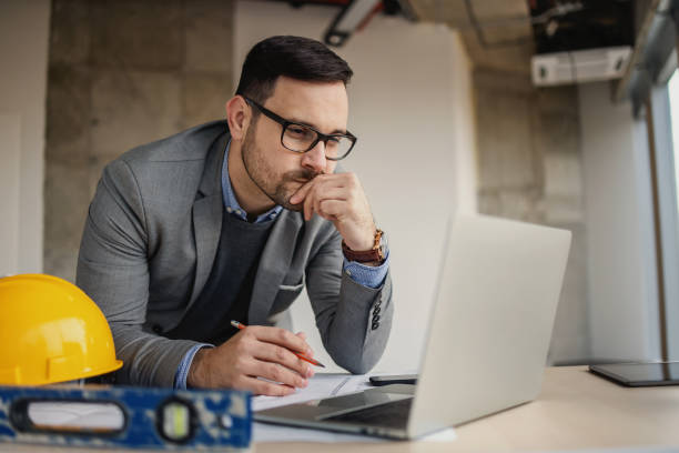 istock Focused architect leaning on desk at construction site with pencil in his hand and looking at laptop. 1300336059