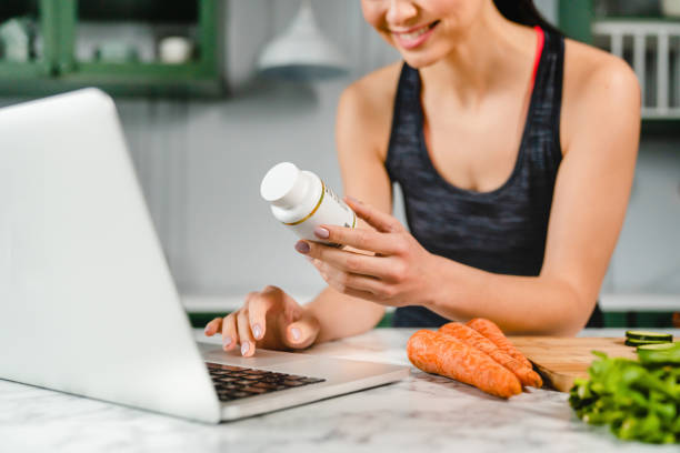 informations heureuses de recherche de jeune femme dans l’ordinateur portatif au sujet des suppléments alimentaires dans la cuisine - food supplement photos et images de collection