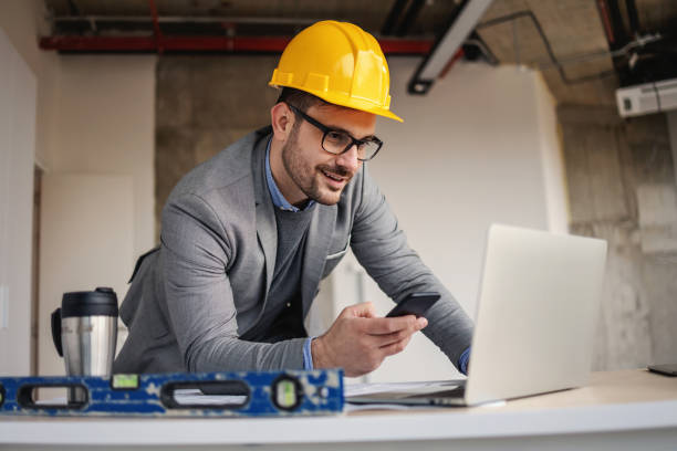 Smiling architect leaning on desk at construction site, looking at laptop and using phone to tell colleagues about project. Smiling architect leaning on desk at construction site, looking at laptop and using phone to tell colleagues about project. lean construction management stock pictures, royalty-free photos & images