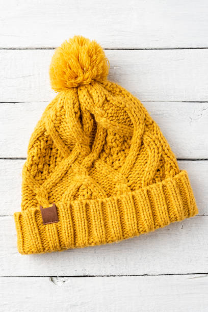 istock Yellow winter hat with pompon on white wooden background. Top view 1300336760