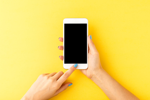 Female hand with blue nails using smart phone with empty screen on yellow background. Top view