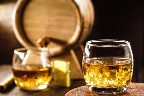 whiskey glass with ice, spot focus, bottle, lighter and barrel on blurred background. distilled drink with malt - cigar whisky bar cognac imagens e fotografias de stock