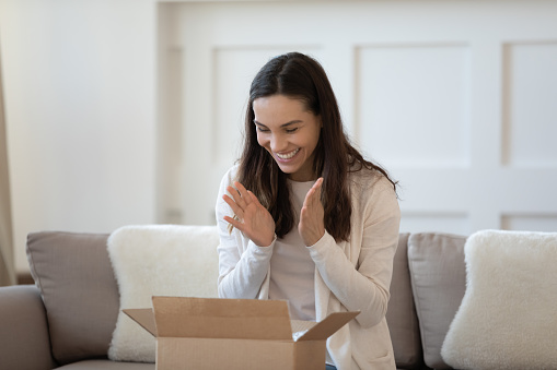 What a cute thing! Excited overjoyed young woman shopper unboxing parcel shipped by mail feeling happy satisfied by quick reliable courier service, high quality of goods products ordered by internet