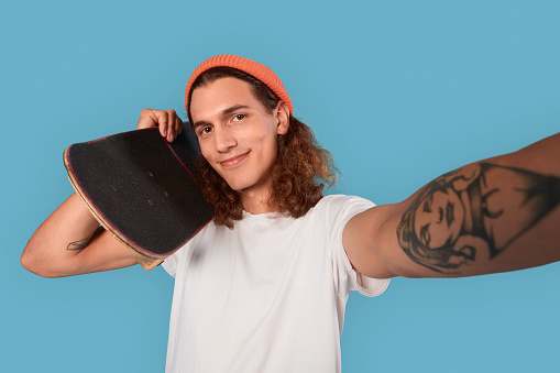 Cheerful and smiling young adult guy in casual wear clothes standing at studio on blue background, holding skateboard, making selfie