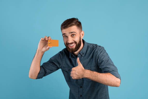 istock Cheerful young guy with credit card showing thumb up gesture on blue studio background 1300345368