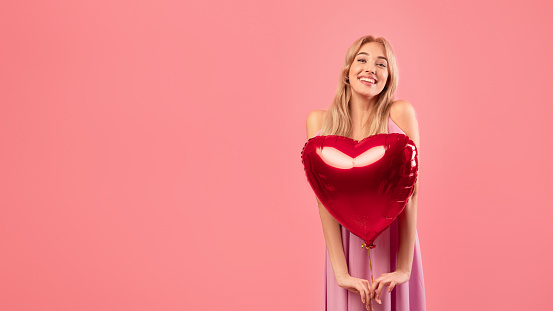 Portrait of happy blonde woman holding heart shaped balloon for Valentine's Day on pink studio background, panorama with empty space. Carefree millennial lady having romantic party on Lovers holiday