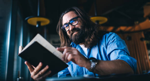 smiling mature man in with long brunette hair spending free time on interesting literature hobby, positive caucasian male 40 years turn pages in book satisfied with leisure and self education in cafe - 40 44 years imagens e fotografias de stock