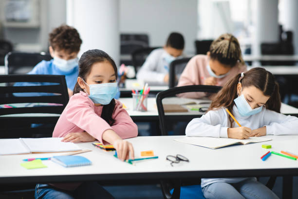 istock Children in masks sitting at desk in classroom 1300351273