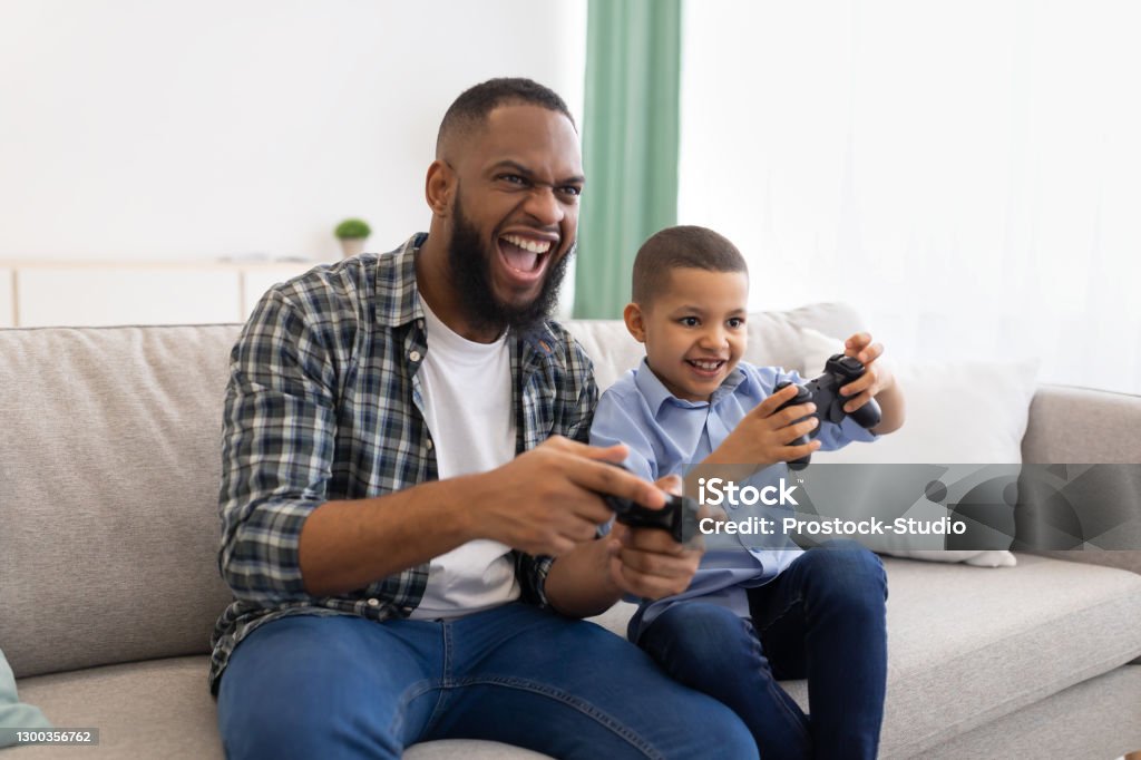 Excited African Dad And Son Playing Game Sitting At Home Excited African Dad And Son Playing Game Having Fun Sitting On Couch At Home. Young Black Father And His Kid Play Videogames Spending Time Together On Weekend Concept Father Stock Photo