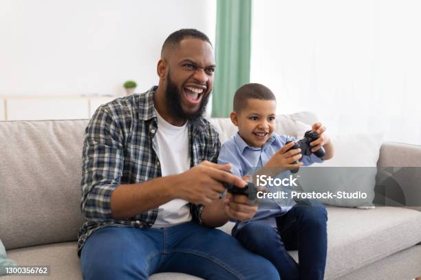 Excited African Dad And Son Playing Game Sitting At Home Stock Photo - Download Image Now