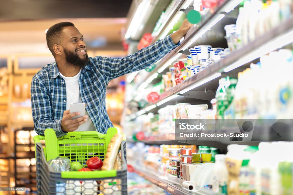 Black Male Shopping Groceries In Supermarket Taking Product From Shelf Black Male Buyer Shopping Groceries In Supermarket Taking Dairy Product From Shelf Standing With Shop Cart Indoors. Guy Buys Grocery Choosing Food In Super Market. Empty Space Supermarket Stock Photo