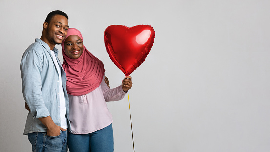 African american muslim couple with love balloon posing cheerfully on grey studio background, panorama with copy space. Merry black lady in hijab hugging her man and holding red heart shaped balloon