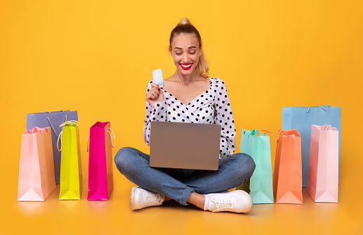 Online Payment Concept. Smiling young woman holding debit credit card and using laptop, ordering clothes and purchases via internet, sitting on the floor with colorful shopping bags at yellow studio