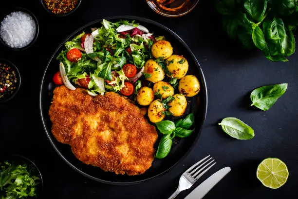 Fried pork chop, potatoes and fresh vegetables on black table