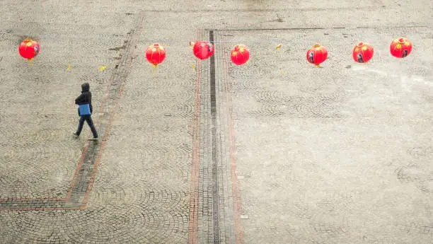 Red chinese lanterns hanging over a tiled square in an urban environment.  A single passerby walks underneath the lanterns towards the left edge of the photo
