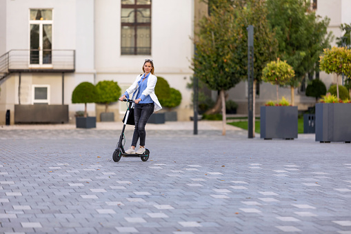 Young Attractive Woman in Business Suit is Riding her Electro Scooter in the City Street and Going to the Office. A Beautiful and Successful Well-Dressed Business Person is Driving his Motorized Scooter Through the City Streets and Rushing to the Work.